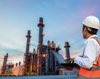 Engineer with notebook standing in front of oil refinery building structure in heavy petrochemical industry